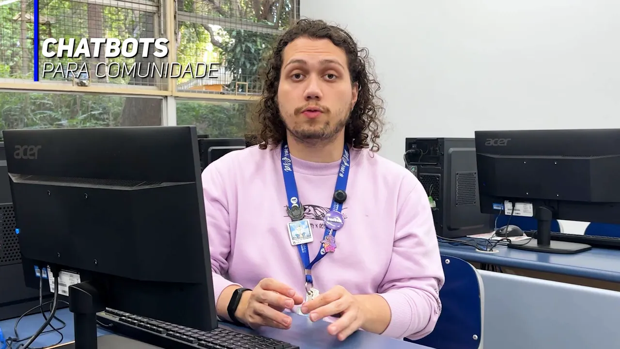 Pessoa em um laboratório de informática com computadores ao redor e selo 'CHATBOTS PARA COMUNIDADE' no canto superior esquerdo, mostrando as mãos em gesto explicativo.